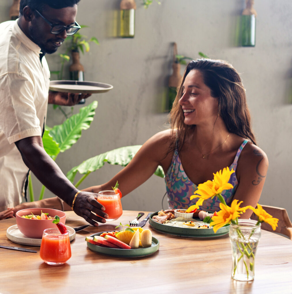 A waiter serving fresh juice to a guest at a breakfast table set with fruit, eggs and sunflowers